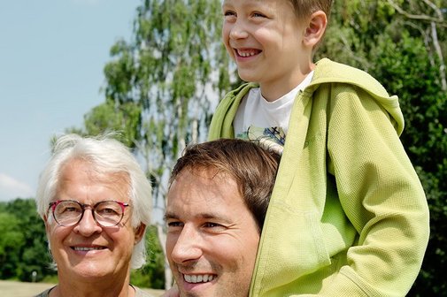 Drei M&auml;nner - ein &auml;lterer Mann l&auml;chelt in die Kamera, ein j&uuml;ngerer Mann blickt in die Ferne, auf seinen Schultern sitzt ein Junge.