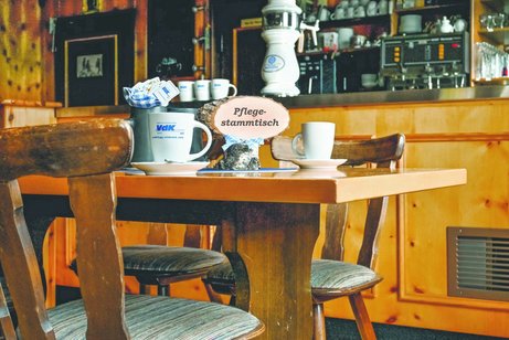 Man sieht den Innenraum einer Wirtschaft. Im Vordergrund sind zwei Stühle und ein Tisch. Auf dem Tisch stehen zwei Tassen und ein Schild mit "Pflegestammtisch".