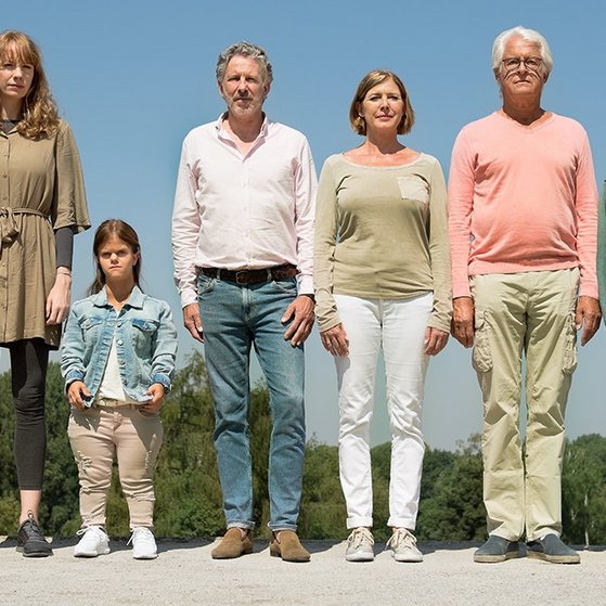 Acht Personen unterschiedlichen Alters und Geschlechts stehen nebeneinander in einer Reihe.