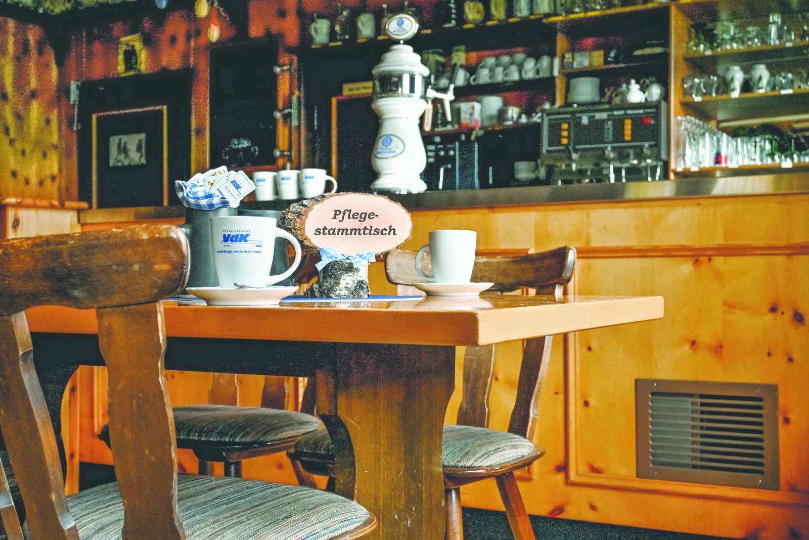 Man sieht den Innenraum einer Wirtschaft. Im Vordergrund sind zwei Stühle und ein Tisch. Auf dem Tisch stehen zwei Tassen und ein Schild mit "Pflegestammtisch".