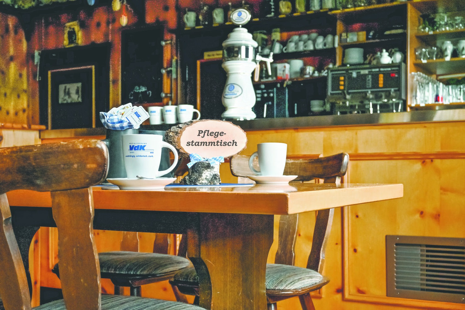 Man sieht den Innenraum einer Wirtschaft. Im Vordergrund sind zwei St&uuml;hle und ein Tisch. Auf dem Tisch stehen zwei Tassen und ein Schild mit "Pflegestammtisch".